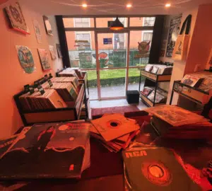 A small record store interior with vinyl records on racks and in stacks, posters on the walls, and a glass door leading to an outdoor area visible in the background.